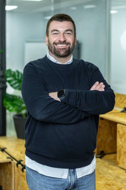 Bir adam kollarını kavuşturarak modern bir çalışma alanında duruyor. Gülümsüyor ve günlük kıyafetler giyiyor. Kurulumda ahşap elementler ve bir bitki yer alıyor.