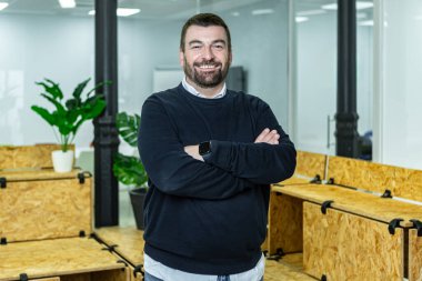 Kolları çapraz bir şekilde çalışma alanında duran bir adam. Bu sette ahşap mobilyalar ve ev bitkileri yer alıyor. Çevre işbirliği ve verimlilik için tasarlanmıştır..