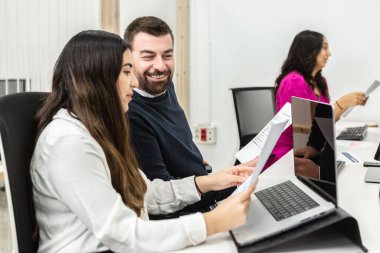 İki profesyonel birlikte çalışıp belgeleri gözden geçirmeye odaklanmış bir çalışma alanı. Parlak, modern ofis ortamında dizüstü bilgisayar ve kağıtlarla donatılmış masalarda oturuyorlar..