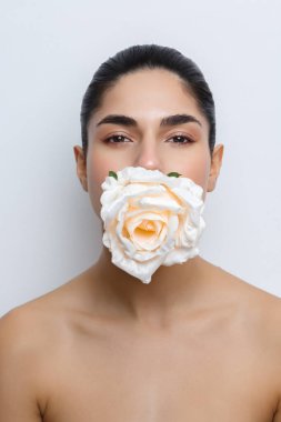 Tarafsız bir arkaplana karşı poz veren bir model, ağzının yanında bir çiçek tutuyor. Odak noktası makyaj ve çiçek aksanları, güzelliği ve yaratıcılığı vurgulamak..