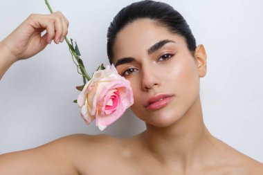 Genç bir kadın stüdyo ortamında yüzünün yanında bir gülle poz veriyor. Doğal güzelliğine ve makyajına odaklanarak, resimdeki çiçek unsurunu vurguluyor..