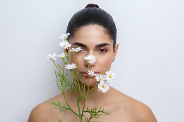 Yüzünün önünde çiçeklerle poz veren bir kadın makyajını renklendiriyor. Ayarlar sade bir arkaplan içeriyor.