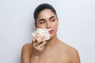 Genç bir kadın yüzüne bir çiçek tutar. Onun doğal bir makyajı ve pürüzsüz bir teni var. Dekor, onun ifadesine ve narin çiçeğine odaklanan tarafsız bir arkaplan içeriyor..