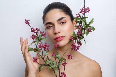 Bir denek, doğal makyaj içeren bir güzellik portresinde poz verir. Tarafsız bir arkaplanda belirlenen çiçeklerle etkileşime geçen kişi..
