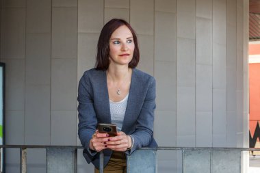 Kısa kızıl saçlı bir kadın elinde telefon olan bir ofisin önünde duruyor. Uzaklara bakarken düşünceli görünüyor..