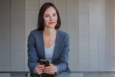 İş kıyafeti giymiş genç bir kadın akıllı telefonuna bakarken korkuluklara yaslanıyor. Sahne, minimalist mimarinin yer aldığı bir şehir ortamında geçiyor..