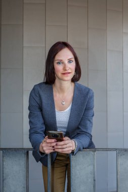 Bir kadın iş elbisesi giyip korkuluklara dayanıyor. O bir akıllı telefon tutuyor ve kameraya bakıyor. Gündüz vakti modern bir bina gibi görünüyor..