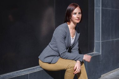 Kısa saçlı genç bir kadın gündüz saatlerinde şehir merkezinde bir çıkıntıda oturur. Basit bir kıyafet giyerken düşünceli ve rahat görünüyor..