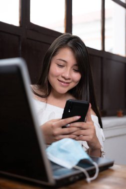 Güzel Latin bir kadın cep telefonunu laptopuyla ve odaktan uzak bir şekilde kullanıyor.