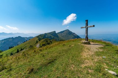 Kırsal ahşap bir haç, nefes kesici dağ manzaraları ve berrak gökyüzü ile çerçevelenmiş yemyeşil bir tepenin üzerinde, Vorarlbergs 'in kalbindeki huzur ve tefekkür manzarasını çağrıştırıyor.