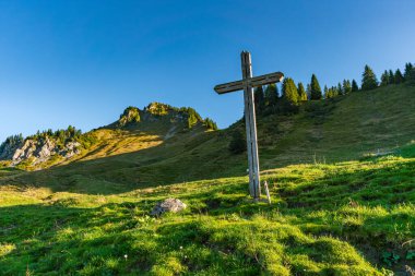 Huzurlu bir ahşap haç, yeşil bir yamaçta gururla duruyor. Avusturya 'nın Alp Dağları' ndaki açık gökyüzünün altında barış ve yansımayı çağrıştırıyor.