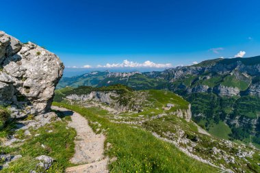 Panoramik bir dağ yürüyüşü geniş alp manzaraları, yemyeşil çayırlar ve açık mavi gökyüzü altında dramatik kaya oluşumlarını gözler önüne seriyor, sükunet ve macera çağrıştırıyor.