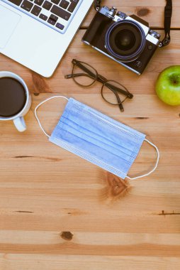 Kamera, dizüstü bilgisayar, gözlük, elma, yüz maskesi ve ahşap arka planda kahve dolu bir kahve fincanı..