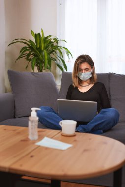 Kanepede bacak bacak bacak üstüne atarak otururken dizüstü bilgisayarıyla çalışan beyaz beyaz bir kız. Tahta masada bir fincan kahve, el dezenfektanı ve yüz maskesi var..