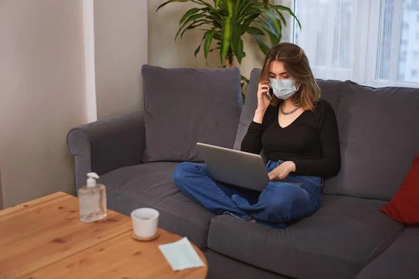 Caucasian white Teenager girl with a facial mask, using computer and talking with her smartphone while sitting on the couch.  There is a coffee cup,  hand sanitizer and a mask on the table.