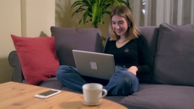 Genç Türk kadın kanepede otururken bilgisayarında bir video görüşmesi yapıyor. Ahşap masada bir kahve fincanı ve akıllı telefon var..