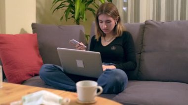 Genç Türk kadın, kanepede otururken akıllı telefonuyla mesajlaşıyor. Masada kahve fincanı, el dezenfektanı ve maske var..