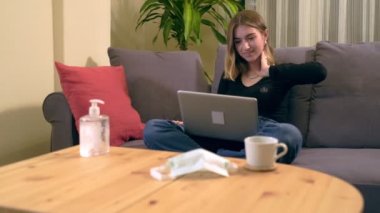 Genç Türk kadın kanepede otururken bilgisayarında bir video görüşmesi yapıyor. Ahşap masada bir kahve fincanı, bir akıllı telefon ve tıbbi maske var..