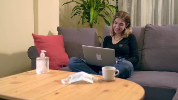 ソファに座っている間 若いトルコ人女性がコンピュータ上でビデオ通話をしています 木製のテーブルにはコーヒーカップ スマートフォンと医療用マスクがあります — ストック動画