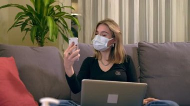 Yüz maskeli genç bir Türk kadın, kanepede otururken telefonuyla video görüşmesi yapıyor. Kucağında dizüstü bilgisayar, kahve fincanı ve el dezenfektanı şişesi var..
