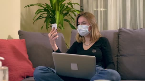 年轻的土耳其女人 戴着面具 坐在沙发上 用智能手机打了一个视频电话 她腿上有台笔记本电脑 桌上有咖啡杯 桌上还有个洗手水壶 — 图库视频影像