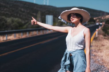 20 'li yaşlarda hasır şapkalı bir kadın yol kenarında otostop çekiyor.