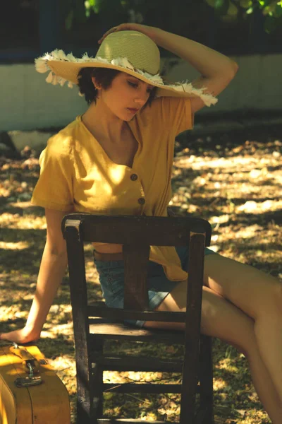 Country woman sits on a wooden chair with a straw hat yellow shirt and jean shorts.