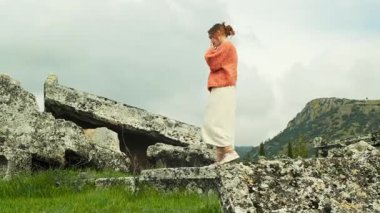 El yapımı turuncu hırka giyen genç bir kadın Hierapolis, Pamukkale 'deki antik harabelerin önünde zarifçe poz veriyor ve hareket ediyor..