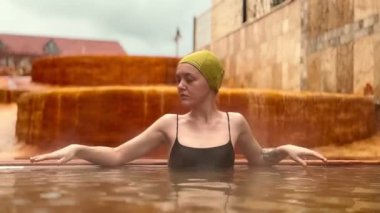 Slav özelliklerine sahip yirmili yaşlarında bir kadın bulutlu bir gökyüzünün altında termal havuzun kenarında dinleniyor..