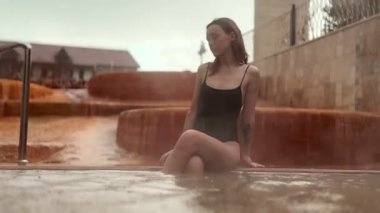 Slav özelliklerine sahip yirmili yaşlarında bir kadın bulutlu bir gökyüzünün altında termal havuzun kenarında dinleniyor..