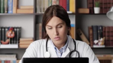 Bilgisayarda çalışan yorgun doktor kahve içiyor. Ofisteki beyaz önlüklü kadın doktor kahve makinesinden mavi bardağa kahve ve içkileri bilgisayar başında çalışırken dolduruyor.