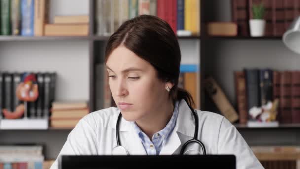 Doctora que trabaja en la computadora, ingresando datos del archivo del paciente. Doctora en bata blanca en su lugar de trabajo en la oficina del hospital trabaja en la computadora, los tipos en el teclado portátil — Vídeos de Stock