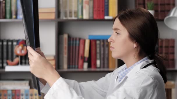 Ο γιατρός εξετάζει ακτινογραφία ή μαγνητική τομογραφία. Συγκεντρωμένη γυναίκα γιατρός με λευκό παλτό στο χώρο εργασίας στο γραφείο διαβάζει μαγνητική τομογραφία ή ακτινογραφία εικόνα και την εξετάζει, κρατά σημειώσεις στο φάκελο του ασθενούς — Αρχείο Βίντεο