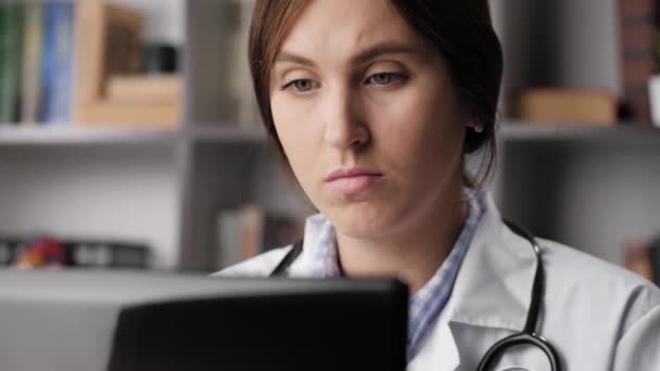 El doctor trabaja en la computadora y bebe café o té. Mujer médico en bata blanca en su lugar de trabajo en la oficina del hospital trabaja en el ordenador portátil, tipos en el teclado y bebe café o té de la taza azul. Primer plano — Vídeos de Stock