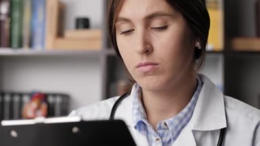 Doktor hasta dosyasını okur ve inceler. Ofisteki beyaz önlüklü kadın doktorun yakın çekimi elinde belgeleri tutuyor ve dikkatlice inceliyor, sayfayı çeviriyor.