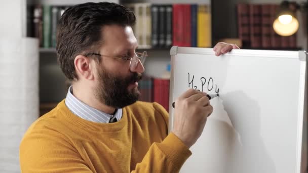 Online-Bildung und Fernlernkonzept. Männlicher Chemielehrer spricht und schaut in die Kamera und schreibt Phosphorsäureformel auf weißes magnetisches Whiteboard, während er der Kamera erklärt — Stockvideo