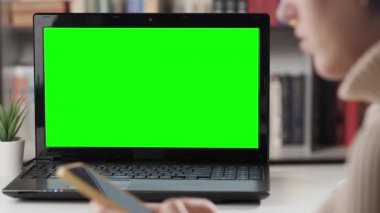 Uzaktan öğrenme dersi ya da webinar, video çağrısı, online eğitim konsepti. Masadaki dizüstü bilgisayarın yakın çekimi, kadın telefonu elinde tutuyor ve akıllı telefondan notlar alıyor, bazen bilgisayara bakıyor ve konuşuyor.