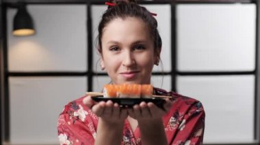 Suşi ikram eden kadın. Asya tarzı giyinmiş çekici bir kadın ve Asya 'nın arka planında Japon usulü suşi tabağı tutuyor ve kameraya sunuyor. Yavaş çekim