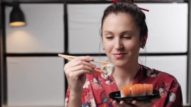 Suşi yiyen bir kadın. Asya tarzı kırmızı elbiseli mutlu bir kadın siyah porselen tabaktan suşi yiyor, yemekten hoşlanıyor. Boşluk bileşimini kopyala