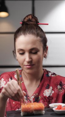 Suşi yiyen bir kadın. Asya tarzı kırmızı elbiseli mutlu bir kadının siyah porselen tabaktan suşi yerken çekilmiş dikey videosu, yemekten hoşlanıyor. Yavaş çekim