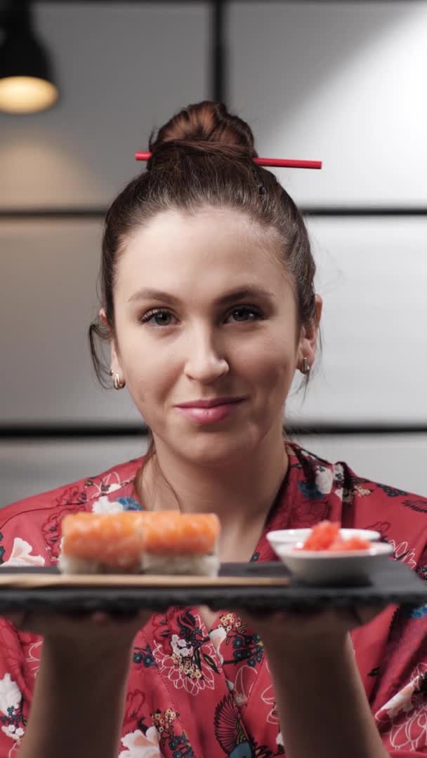 Woman offering sushi. Vertical video of smiling woman dressed in Asian style and against background of Asian Japanese interior holds plate of sushi rolls in her hands and offers them to camera — Stock Video