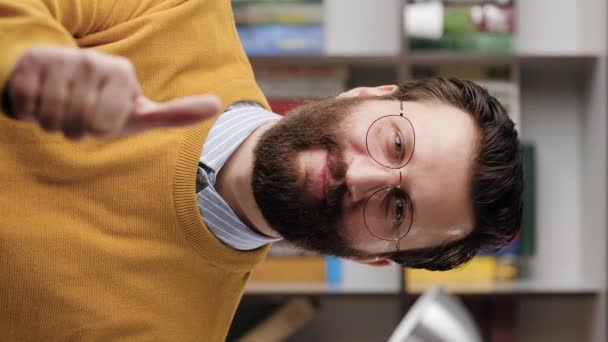 Levez le pouce. Vidéo verticale de l'homme barbu souriant positif dans des lunettes dans le bureau ou la pièce de l'appartement en regardant la caméra et montre son pouce vers le haut. Gros plan et ralenti — Video
