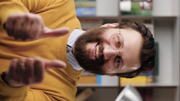Άντρας με το δάχτυλο. Κατακόρυφο βίντεο του θετικού γενειοφόρου άντρα με γυαλιά στο γραφείο ή στο δωμάτιο του διαμερίσματος κοιτάζοντας την κάμερα και δείχνει τα δύο χέρια του πάνω. Κοντινό πλάνο και αργή κίνηση — Αρχείο Βίντεο