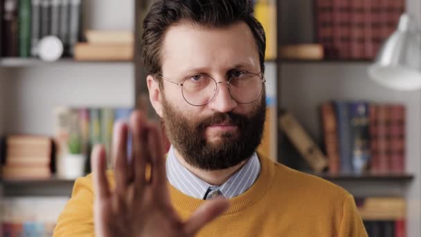 Man show STOP, NO. Serious displeased bearded man in glasses in office or apartment looking at camera and shows his palm STOP. Close-up and slow motion — Wideo stockowe