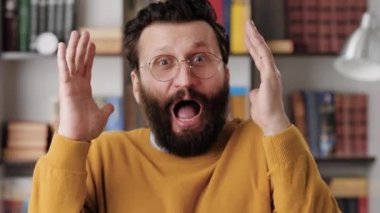 Man is scared, terrified. Frightened bearded man in glasses in office or room in apartment with suddenly throws his hands up and looking at camera, he is very scared and just terrified. Close-up