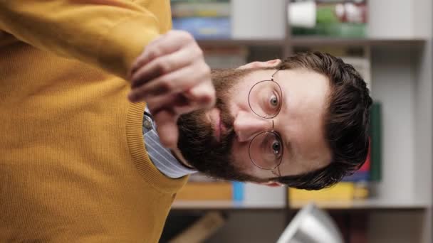 Han vinkar med fingret. Vertikal video av allvarlig rynkar pannan skäggig man med glasögon i kontor eller lägenhet rum tittar på kameran och pekar fingrarna gester till honom. Närbild och slow motion — Stockvideo