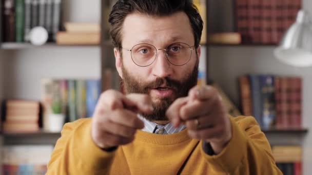 Мужчина указывает на тебя пальцем. Серьёзный нахмуренный бородатый мужчина в очках в офисе или в квартире смотрит в камеру, показывает пальцем и говорит ВЫ. Крупный план и замедленная съемка — стоковое видео