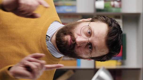 L'homme pointe son doigt vers toi. Vidéo verticale d'un homme barbu froncant les sourcils avec des lunettes dans un bureau ou un appartement regardant la caméra et pointant du doigt et disant VOUS. Gros plan et ralenti — Video
