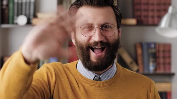 Homem acenando HELLO. Sorrindo positivo rindo homem no escritório ou sala de apartamento olhando para a câmera e acenando mão direita mostrando e diz saudação HI. Close-up e câmera lenta — Vídeo de Stock