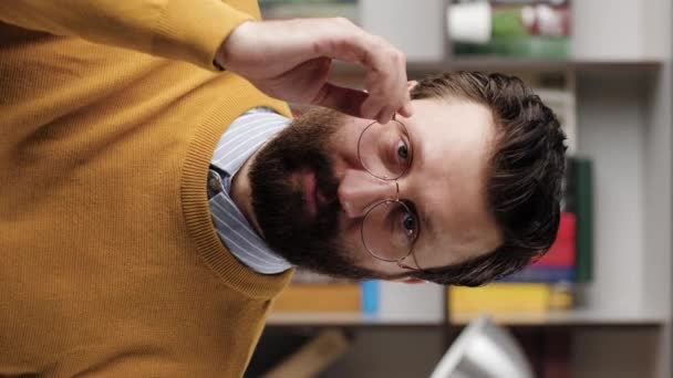 Un clin d'œil. Vidéo verticale de l'homme barbu souriant positif avec des lunettes dans le bureau ou la pièce de l'appartement en regardant la caméra et abaisse lentement ses lunettes et ses clins d'oeil. Gros plan et ralenti — Video